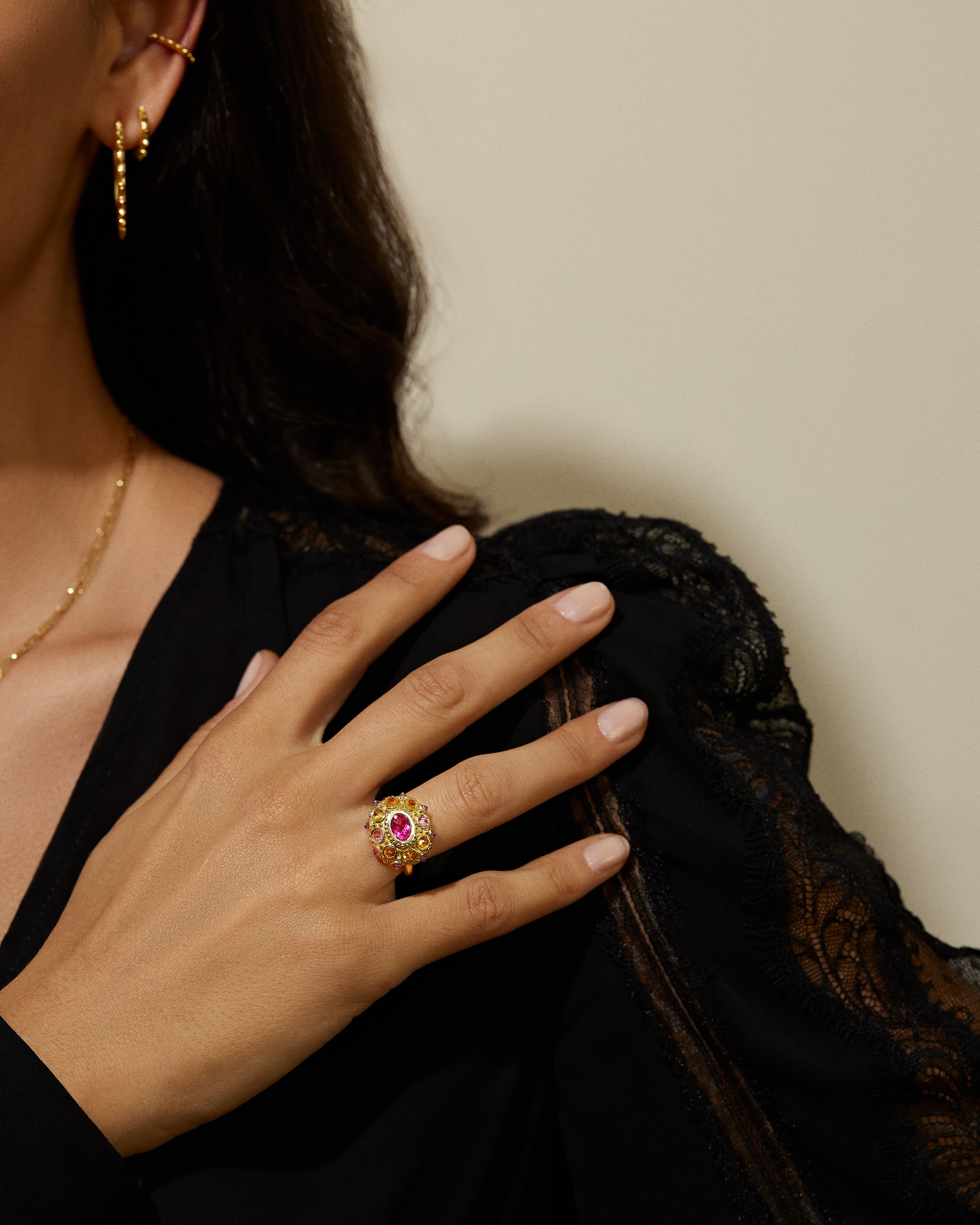 A model in a black blouse showcasing an exquisite 18K yellow gold and precious gemstone ring designed by AnaKatarina Fine Jewelry. Adorned with inverted Rhodalite Garnets, Pink Sapphires, and Orange Sapphires this playfully stunning ring features a large, captivating diamond-cut pink sapphire in the center as its focal point.