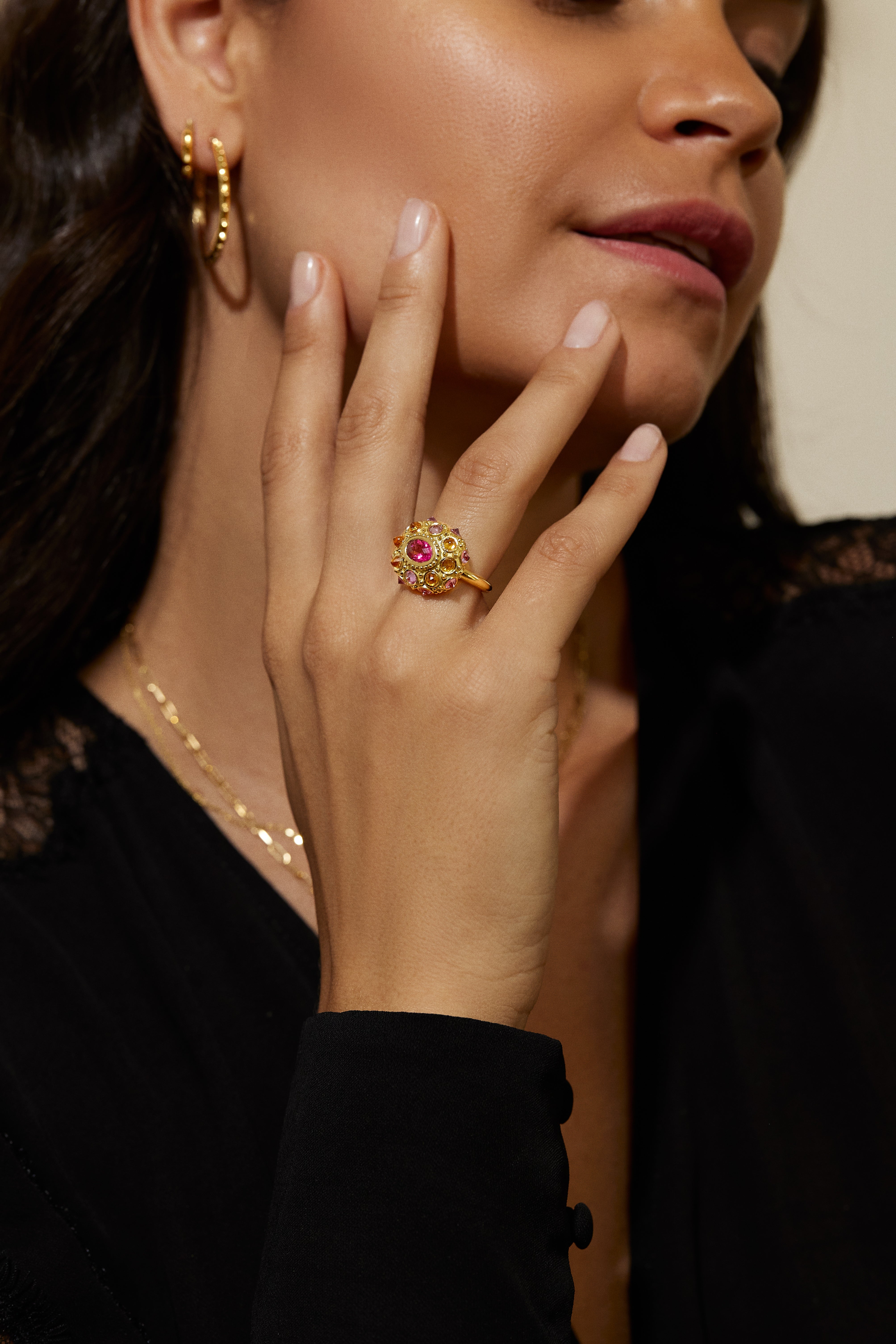 A model in a black blouse wearing an exquisite 18K yellow gold and precious gemstone ring designed by AnaKatarina Fine Jewelry. Adorned with inverted Rhodalite Garnets, Pink Sapphires, and Orange Sapphires this playfully stunning ring features a large, captivating diamond-cut pink sapphire in the center as its focal point.