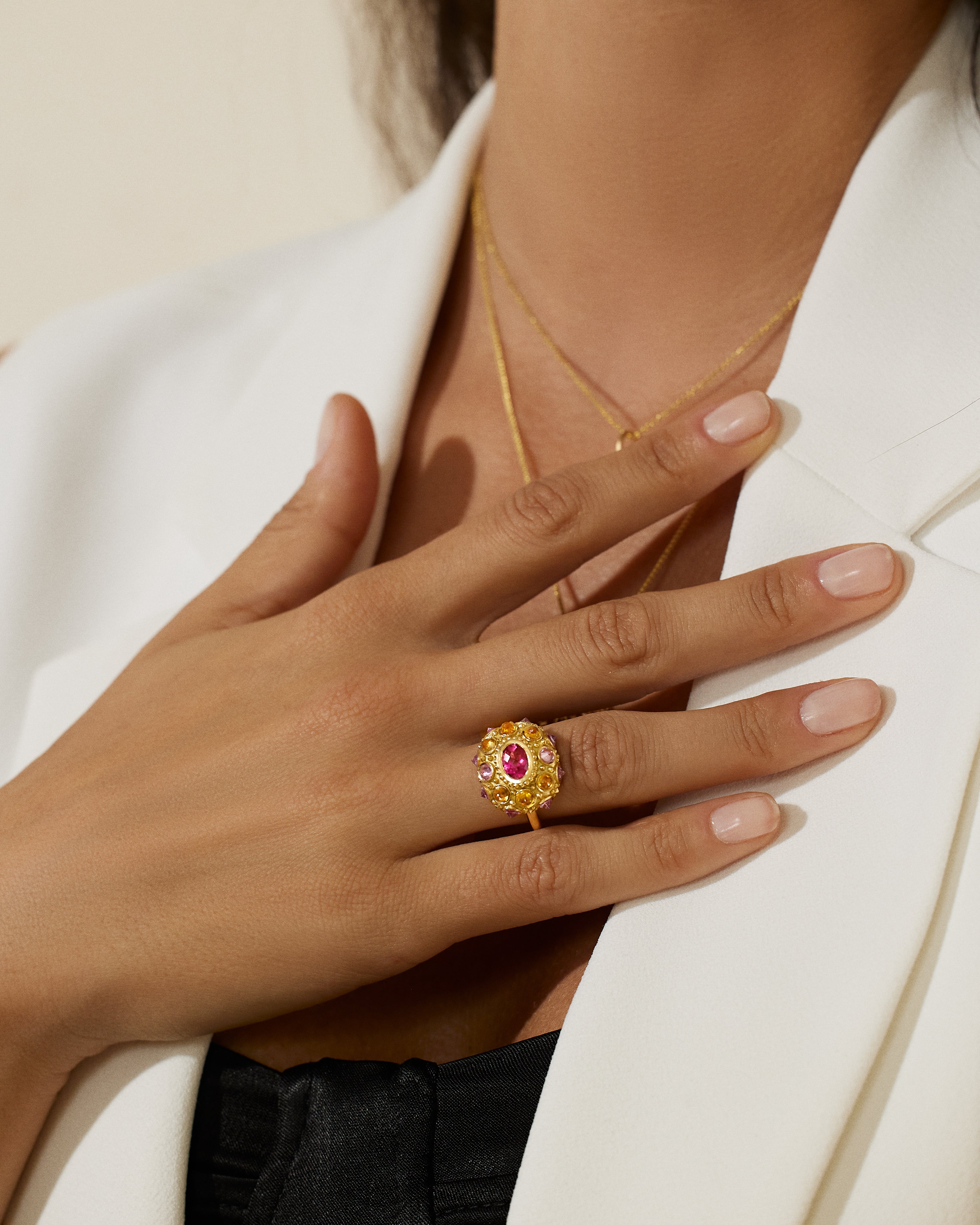 A closeup of a model showcasing an exquisite 18K yellow gold and precious gemstone ring designed by AnaKatarina Fine Jewelry. Adorned with inverted Rhodalite Garnets, Pink Sapphires, and Orange Sapphires this playfully stunning ring features a large, captivating diamond-cut pink sapphire in the center as its focal point.