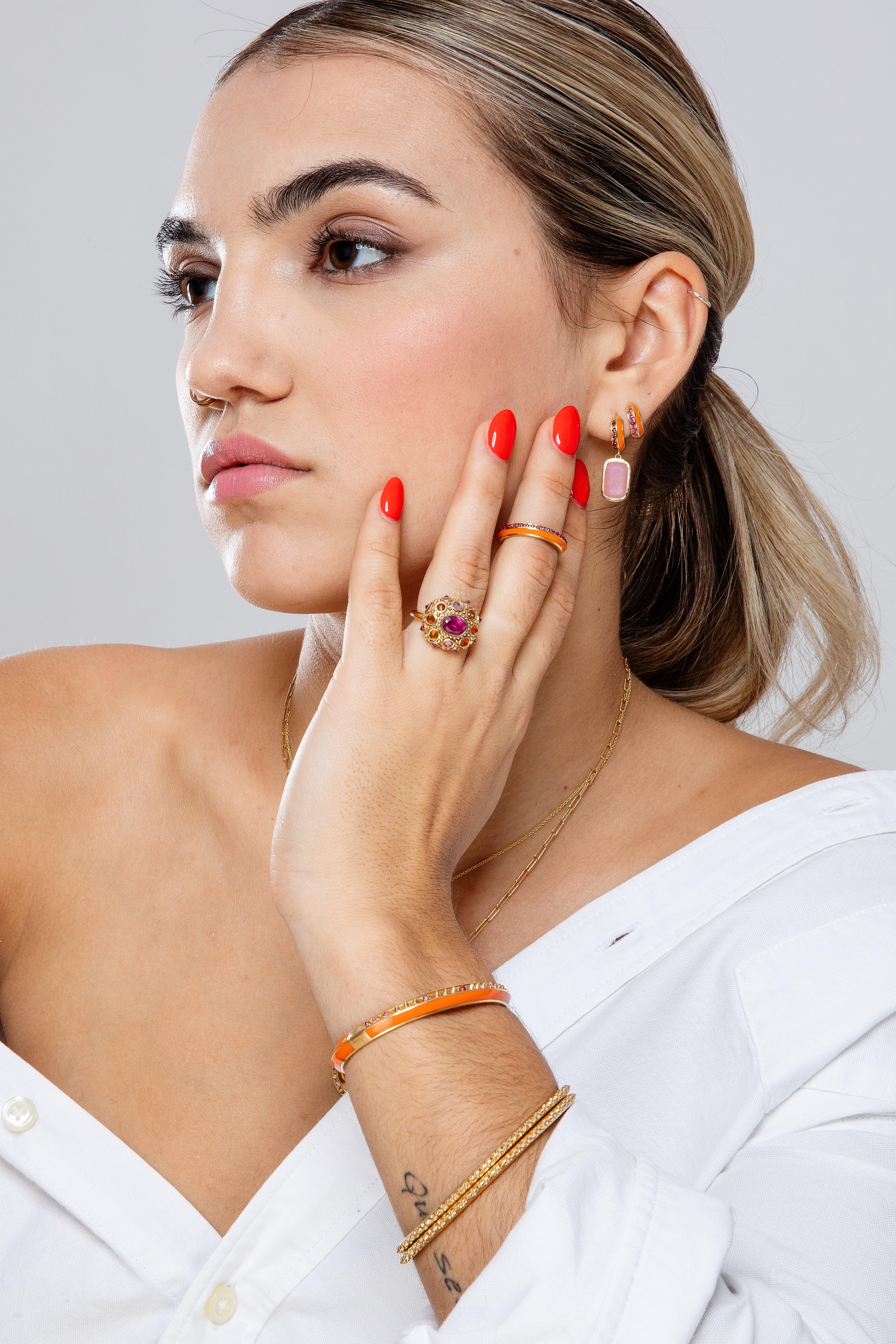 A person modeling 18k gold and pink tourmaline fine jewelry, featuring the 'Sweet, Happy Life' Huggies earrings with .48tw pink tourmaline and orange enamel from Ana-Katarina's Attitude 'Sweet Happy Life' collection and other fine 18k gold chains and bangles.