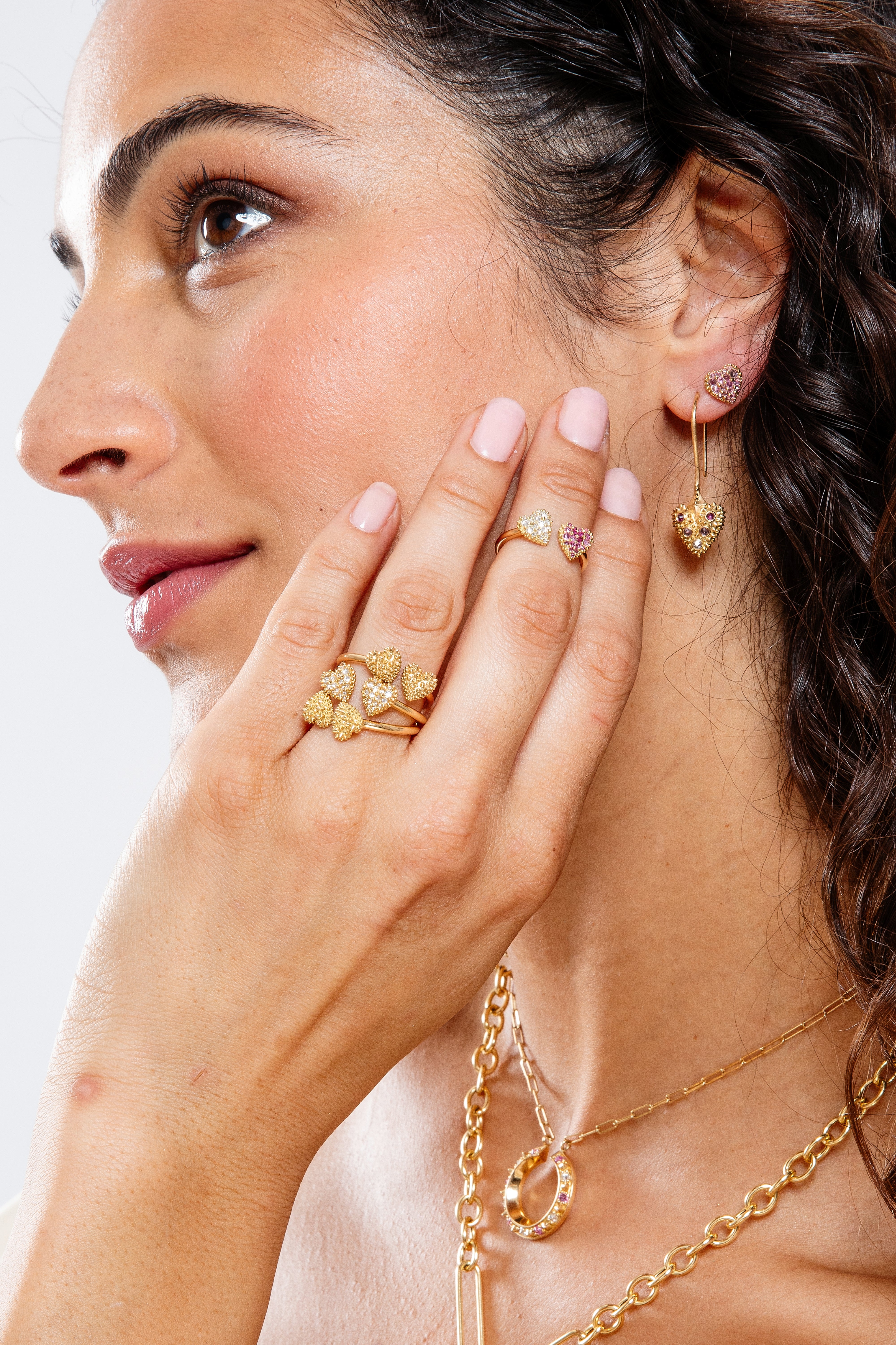 A person modeling variations of an 18k gold & precious gemstone ring with two spiked hearts side by side with pink sapphire heart stud earrings designed by Ana-Katarina fine jewelry. 