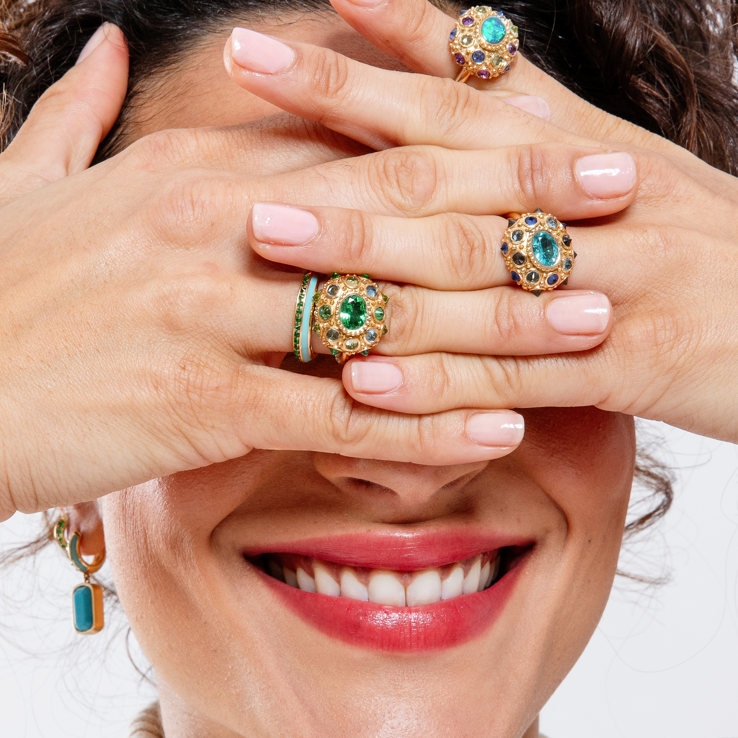 A model smiling & covering her face with her fingers while wearing four rings from Ana-Katarina’s Attitude collection with Tsavorite Garnets, Aquamarines & Peridot in the colors of blue & green creating a captivating & colorful display.