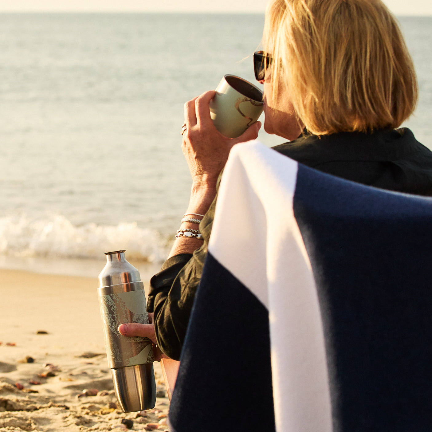 A person with short blond hair sits on a beach chair, drinking from a cup and holding a Caskata x High Camp Light Blue Lucy Parkside Flask by Caskata, facing the ocean.