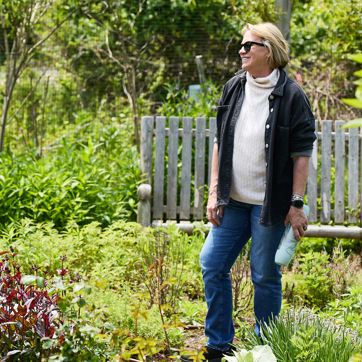 A person in sunglasses and casual clothes stands in a garden, holding the Caskata x High Camp Light Blue Lucy Parkside Flask by Caskata, with a wooden bench and green plants visible in the background.
