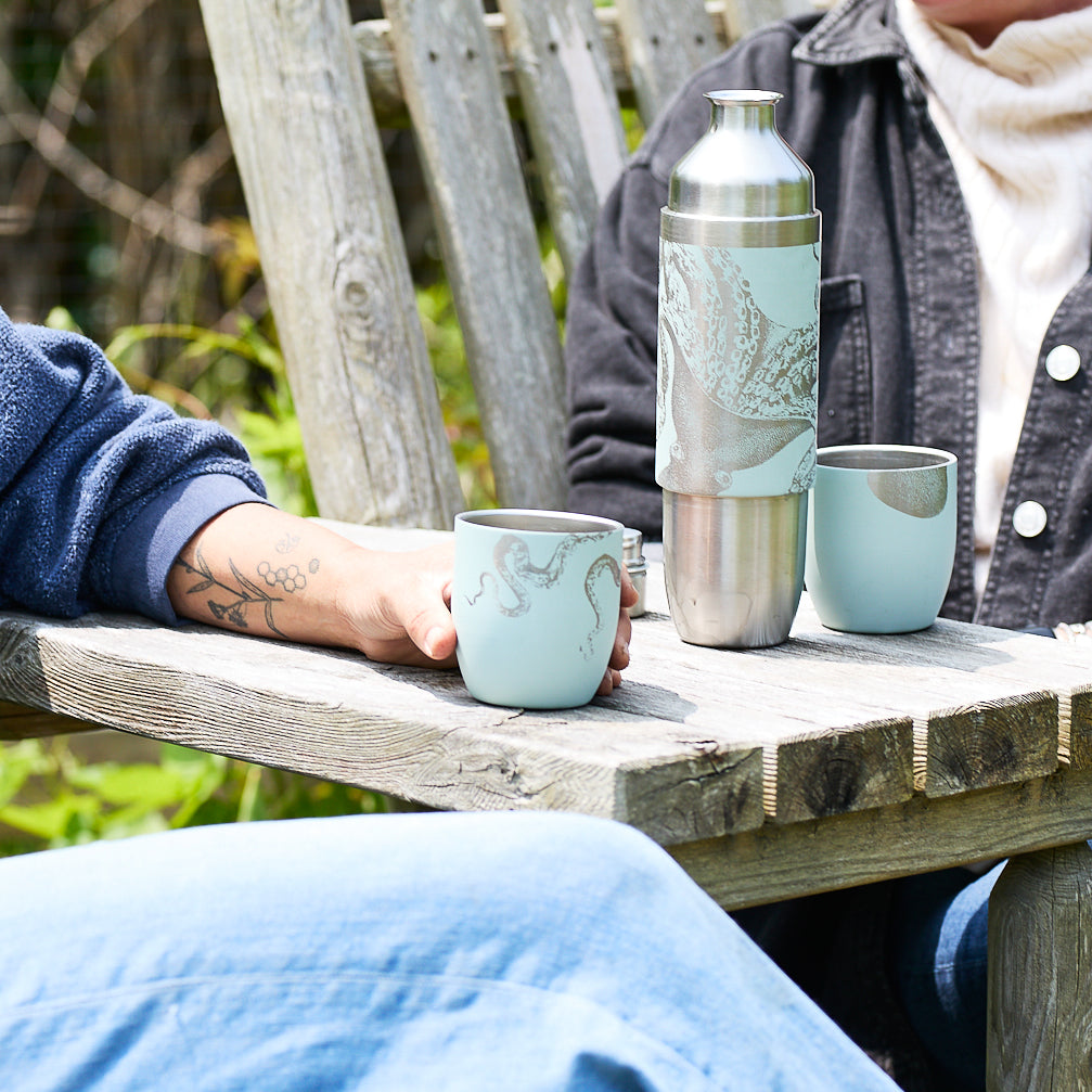 Two people sit outdoors at a wooden table with the Caskata x High Camp Light Blue Lucy Parkside Flask by Caskata and two matching light blue cups featuring an octopus design.