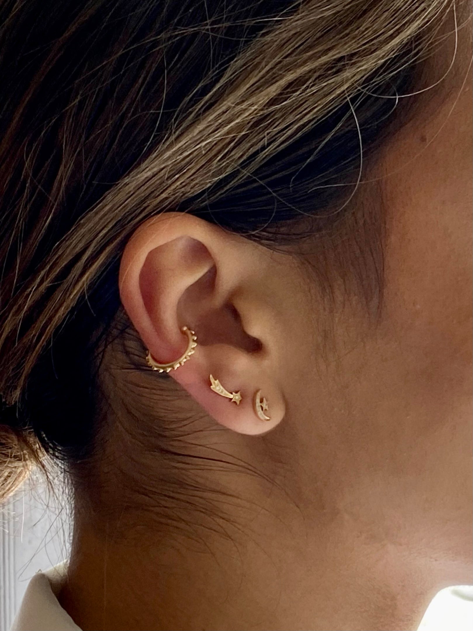 A closeup of a model wearing both 4 Elements: 'Air' stud earrings: the star & moon & the shooting star designs both in 18k yellow gold. Shown with a spiked gold cuff from the Attitude Collection by Ana-Katarina. 