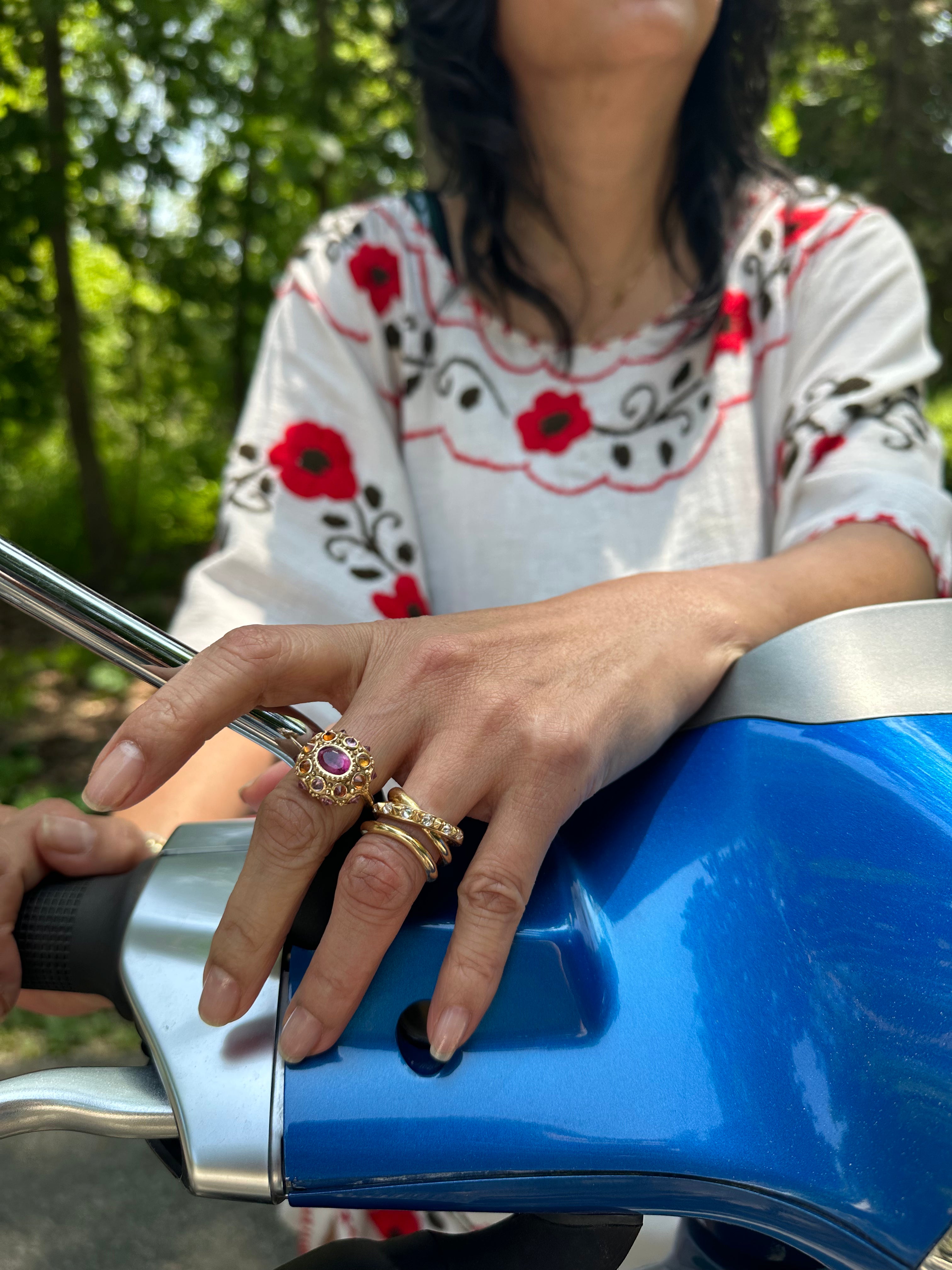 A person with her hand on a blue motorcycle showcasing Ana-Katarina's ‘Amy's Attitude Diamond Ring,’ showcasing elegant, interwoven 18k yellow gold bands embellished with inverted diamonds for a touch of opulence & bold luxury.