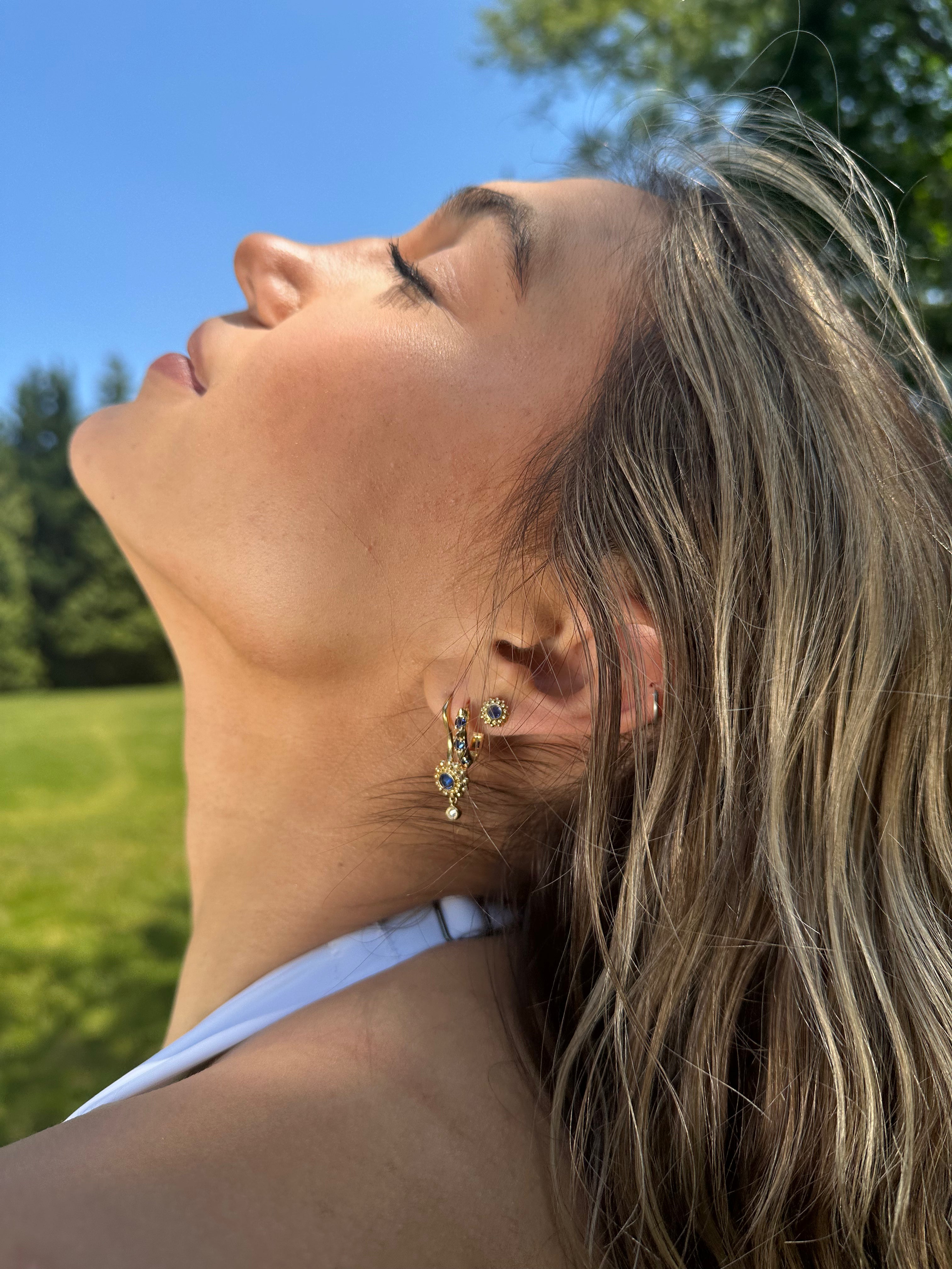 A person modeling various hoops and stud earrings in 18k yellow gold, diamonds, and blue sapphires all designed by Ana-Katarina Fine Jewelry. 