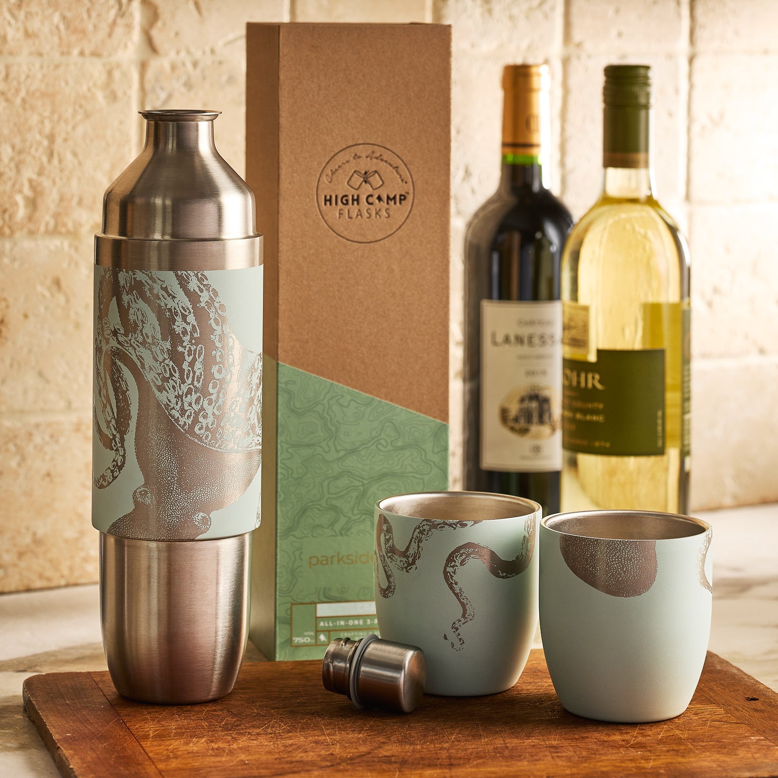 The Caskata x High Camp Light Blue Lucy Parkside Flask is pictured with stainless steel cocktail shaker, two magnetic octopus tumblers, wine bottles, and packaging box arranged on a wooden surface.