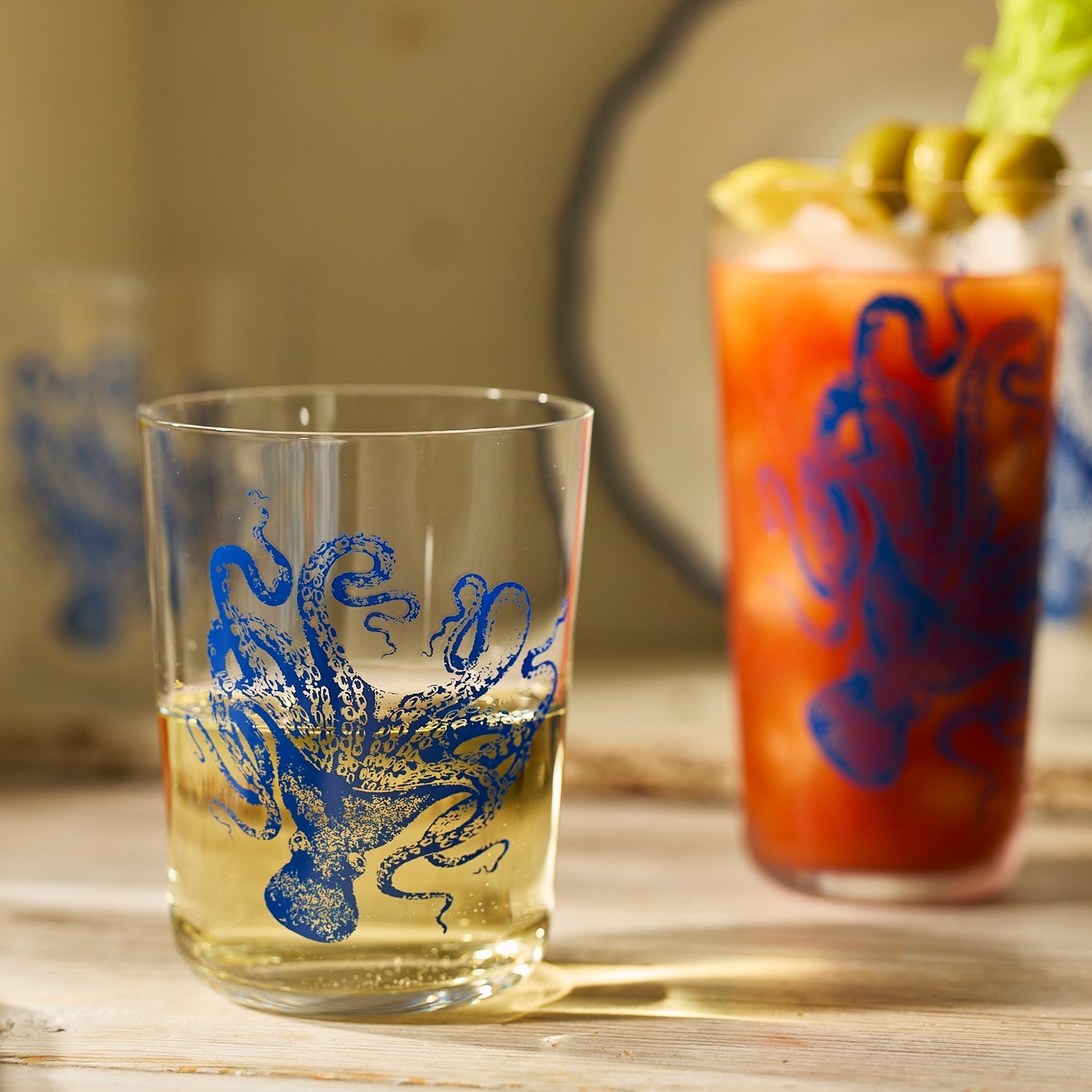 Two Lucy Blues Short Drinking Glasses by Caskata, made from lead-free crystal, feature vivid blue octopus designs—one holds a pale drink, the other a red beverage garnished with olives and celery.