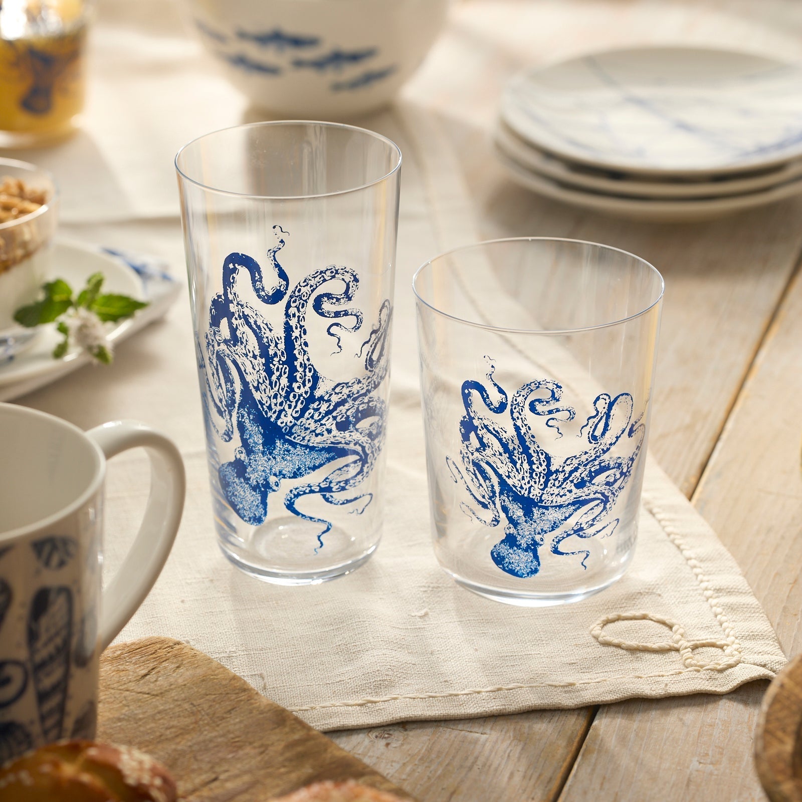 Two Caskata Lucy Blues Short Drinking Glasses with blue octopus designs sit on a cream cloth among dishes and a mug, atop a wooden surface—ideal for those seeking unique, lead-free crystal drinkware.