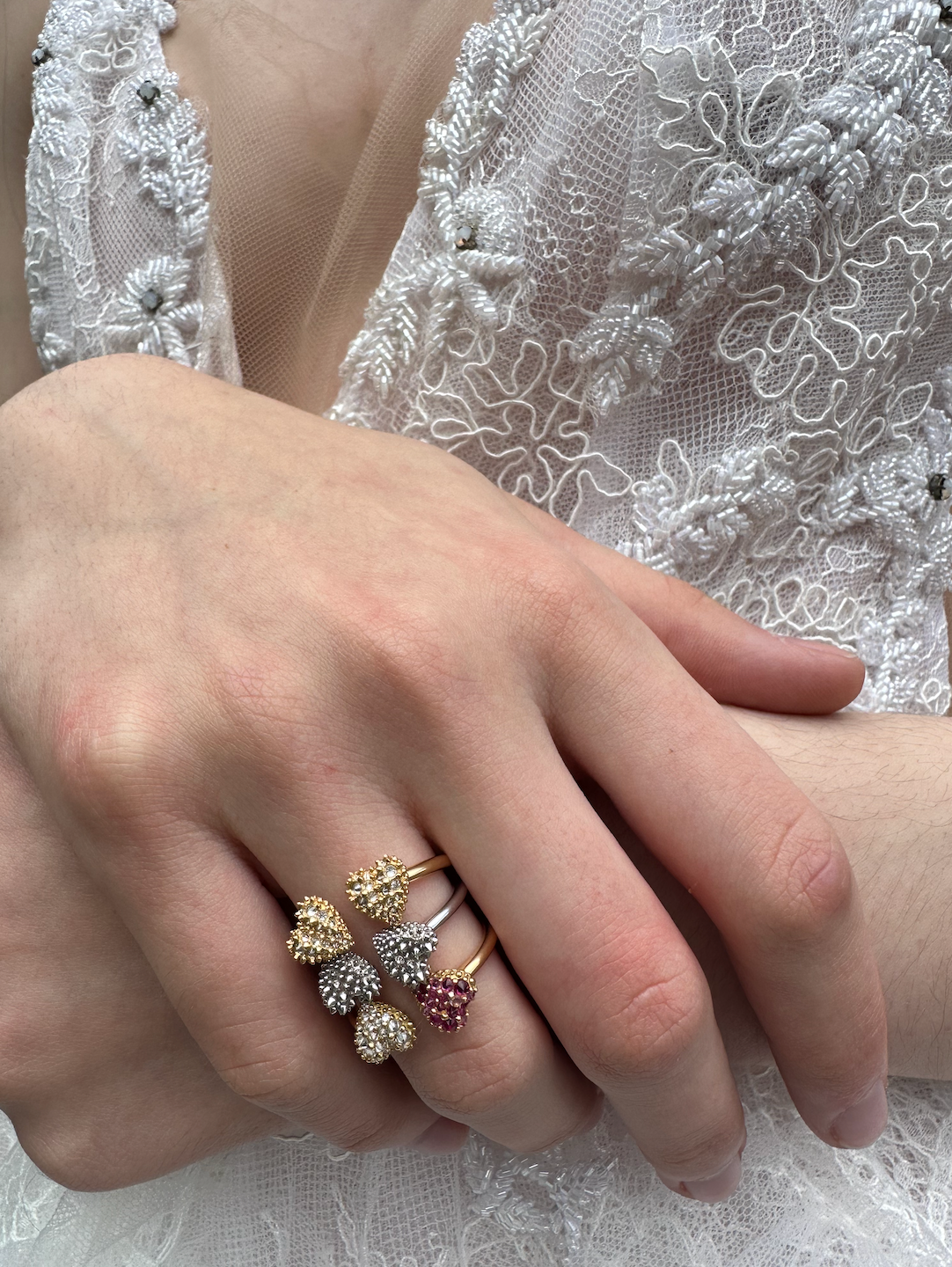 A closeup of a person modeling three variations of the ‘Moi et Toi’ classic ring design in 18k gold & precious gemstones ring with two spiked hearts side by side by Ana-Katarina fine jewelry. 