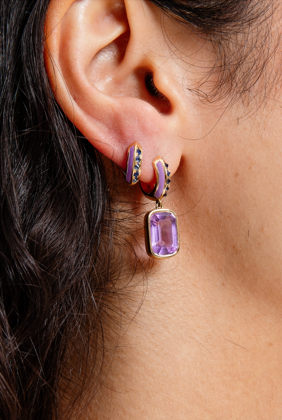 A closeup of a person wearing exquisite drop earrings designed by Ana-Katarina Fine Jewelry in opulent purple and blue hues, featuring .62-carat blue sapphires, 5.3-carat amethyst drop, lavish lavender enamel, and 18k yellow gold.