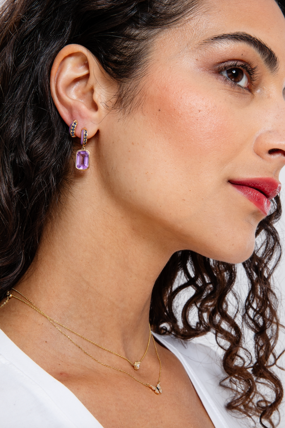 A person wearing exquisite huggie earrings and matching drop earrings designed by Ana-Katarina Fine Jewelry in opulent purple and blue hues, featuring .62-carat blue sapphires, a 5.3-carat amethyst drop, lavish lavender enamel, and 18k yellow gold.