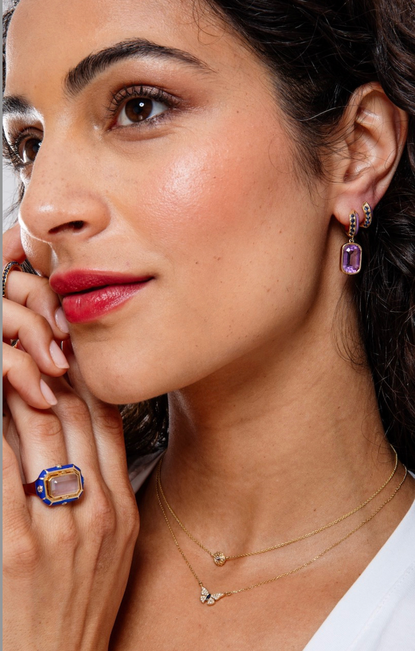 A model wearing two necklaces, earrings, and a poison ring designed by Ana-Katarina Fine Jewelry - both necklaces are in 18k yellow gold with delicate, adjustable 18’ cable chains. One is a diamond solitaire necklace inspired by sea urchins and the other is a diamond and blue sapphire butterfly necklace. 
