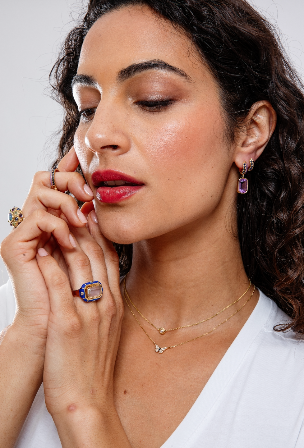 A person modeling exquisite huggie earrings, matching drop earrings, and other fine jewelry designed by Ana-Katarina Fine Jewelry in opulent purple and blue hues. The drop earrings feature .62-carat blue sapphires, a 5.3-carat amethyst drop, lavish lavender enamel, and 18k yellow gold.