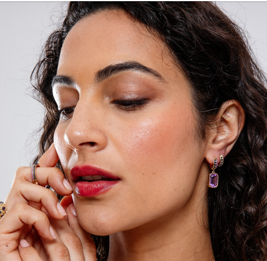 A person with dark curly hair wearing exquisite huggie earrings paired with drop earrings and a matching ring designed by AnaKatarina in opulent purple and blue hues, featuring a .62tw blue sapphires, lavish lavender enamel, and 18k yellow gold.