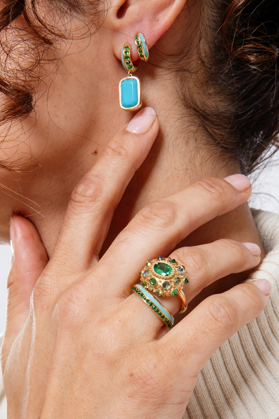 A closeup of a person modeling multiple pieces of tsavorite garnet and turquoise fine jewelry from Ana-Katarina's 'Sweet, Happy Life' collection featuring Tsavorite Garnet Drop Earrings in 18k yellow gold and embellished with .55cttw Tsavorite Garnet, 7.85ct Turquoise and Turquoise Enamel. 