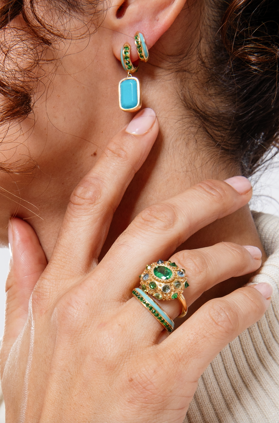 A closeup of a person modeling multiple pieces of tsavorite garnet and turquoise fine jewelry from Ana-Katarina's 'Sweet, Happy Life' collection featuring Tsavorite Garnet Drop Earrings in 18k yellow gold and embellished with .55cttw Tsavorite Garnet, 7.85ct Turquoise and Turquoise Enamel. 