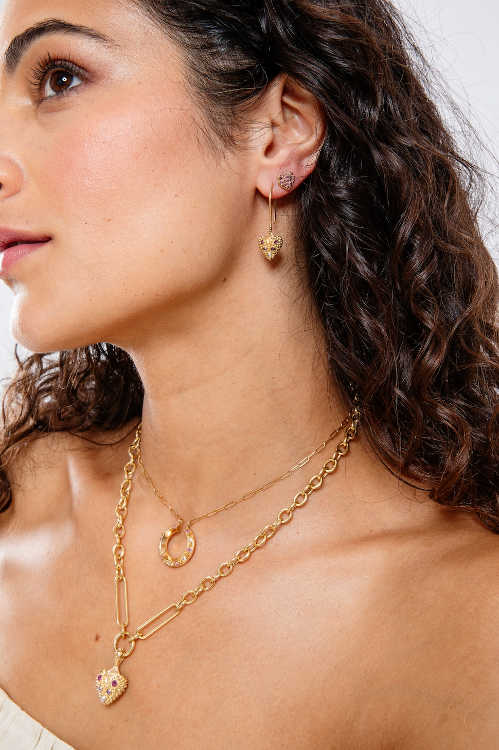 A person modeling 18k gold & inverted diamond & pink sapphire heart drop earrings & stud earrings with other fine jewelry from the ‘Pierce Your Heart’ collection designed by Ana-Katarina Fine Jewelry.