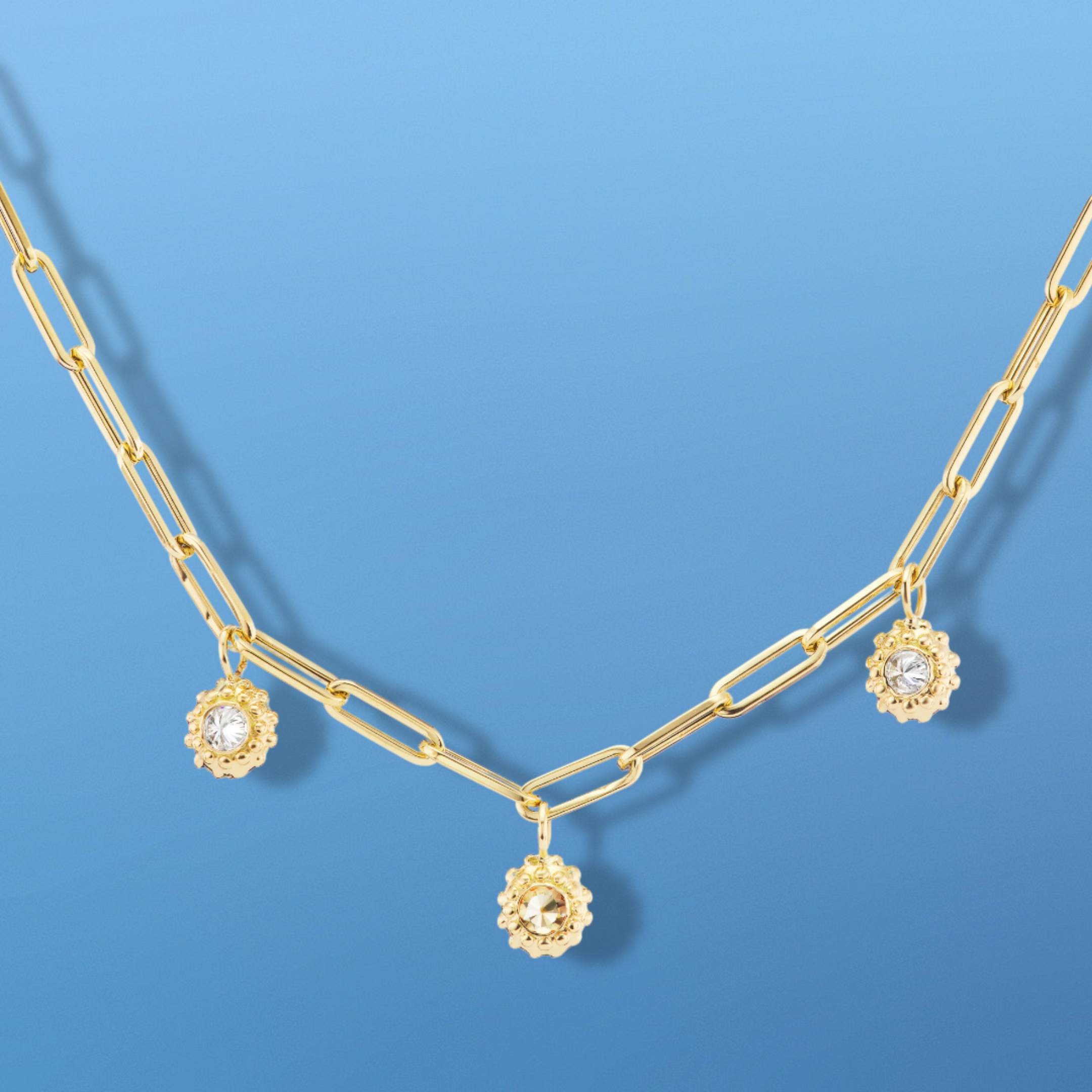 An 18k yellow gold paperclip chain with three ‘Tribe’ diamond solitaire pendants with inverted diamonds and gold beading to resemble sea urchins shown on a aqua blue background. Designed by Ana-Katarina Fine Jewelry. 