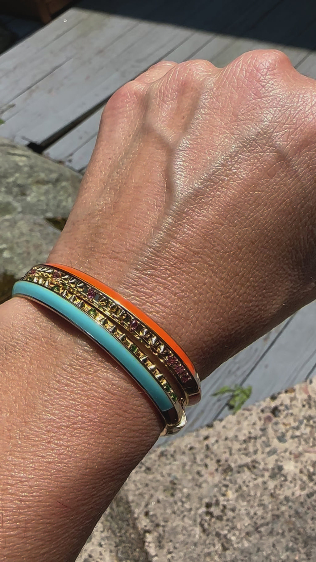 A video of a person modeling two bangle bracelets designed by Ana-Katarina Fine Jewelry stacked together. Both are crafted from 18k yellow gold. One is the 'Sweet, Happy Life' Pink Tourmaline Bangle embellished with .40tw pink tourmaline, gold spikes, and orange enamel. 