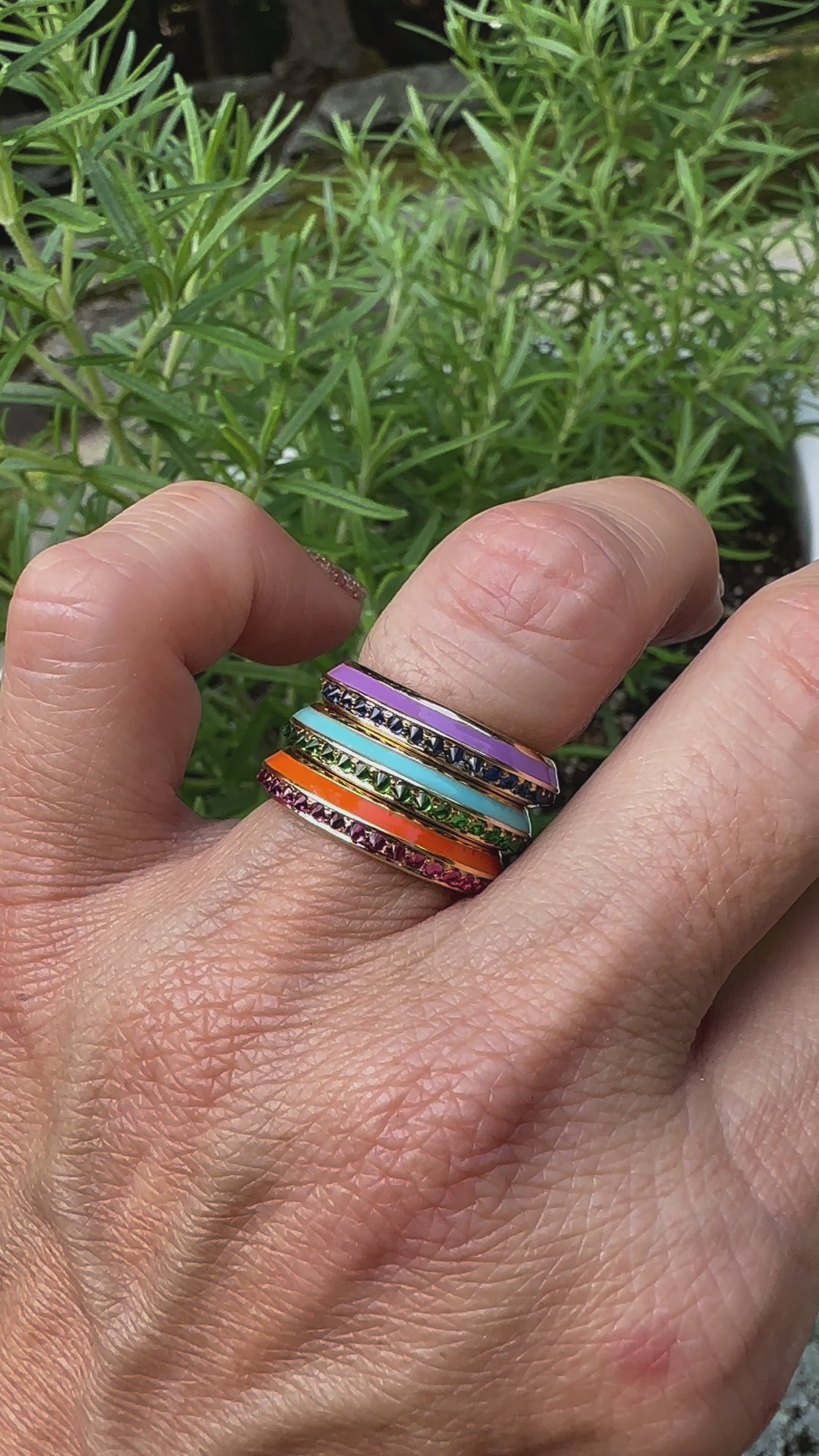 A video of a model wearing three stacking rings from Ana-Katarina's 'Sweet, Happy Life' jewelry collection. Stacked from top to bottom: the 'Sweet, Happy Life' Blue Sapphire Stacking Ring, the 'Sweet, Happy Life' Tsavorite Garnet Stacking Ring, and the 'Sweet, Happy Life' Pink Tourmaline Stacking Ring - all in 18k yellow gold with inverted gemstones and colorful enamel insets. 