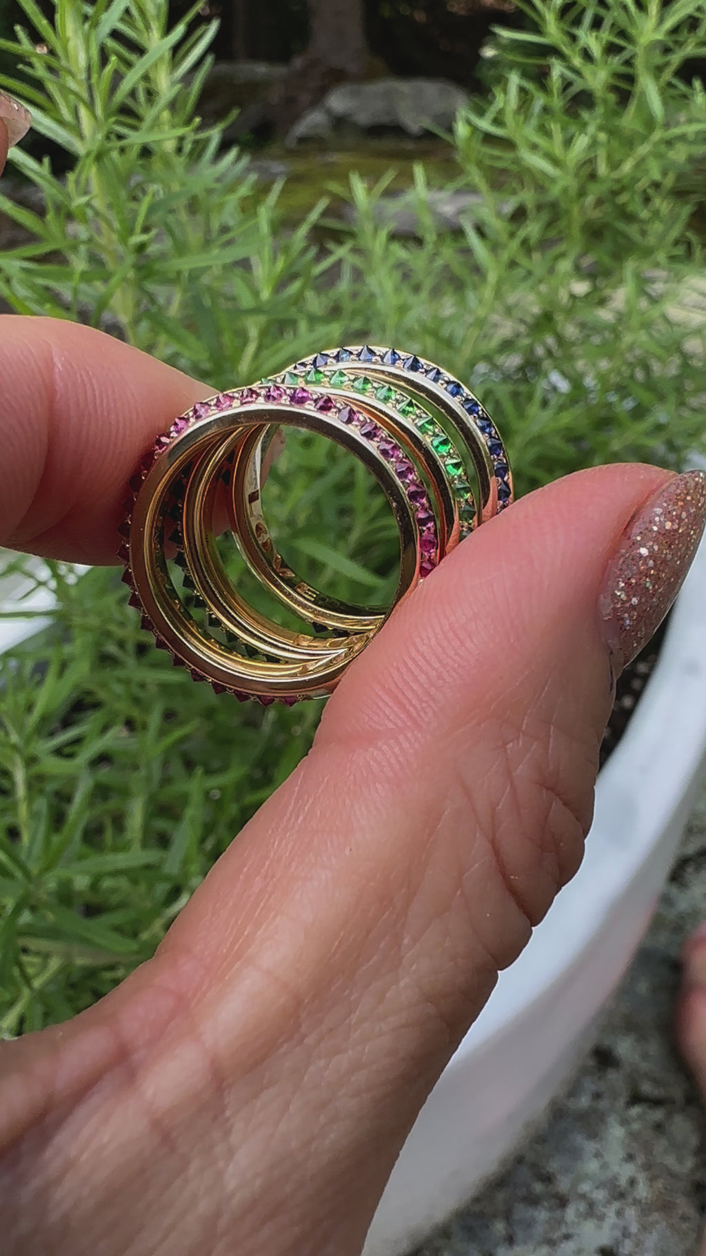 A video of a model holding three stacking rings from Ana-Katarina's 'Sweet, Happy Life' jewelry collection. Stacked from top to bottom: the 'Sweet, Happy Life' Blue Sapphire Stacking Ring, the 'Sweet, Happy Life' Tsavorite Garnet Stacking Ring, and the 'Sweet, Happy Life' Pink Tourmaline Stacking Ring - all in 18k yellow gold with inverted gemstones and colorful enamel insets. 