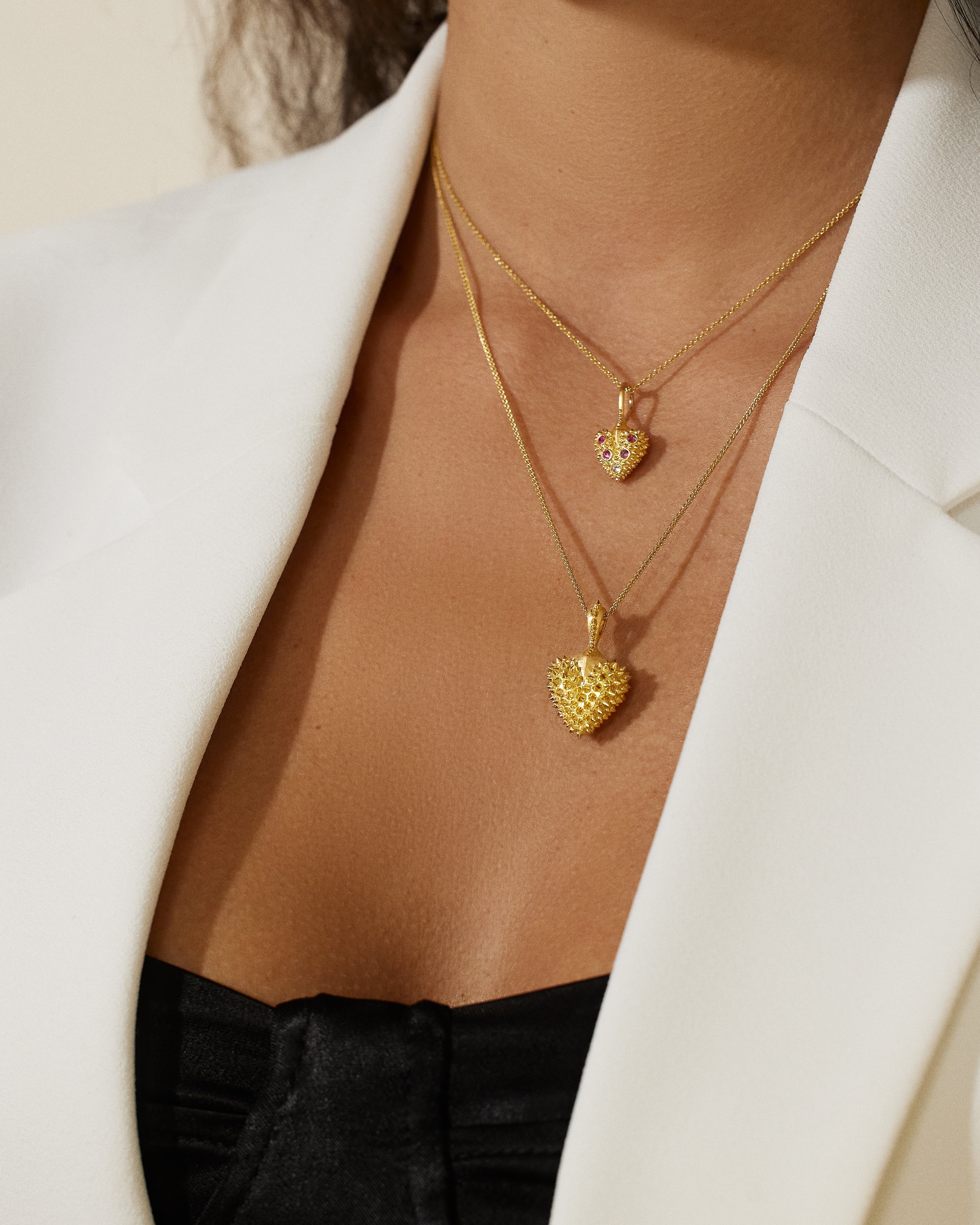 A model wearing Ana-Katarina’s 18k yellow gold heart necklace with the pink sapphire spike heart necklace - both from the ‘Pierce Your Heart’ collection - both on varying lengths of 18k yellow gold cable chains. 