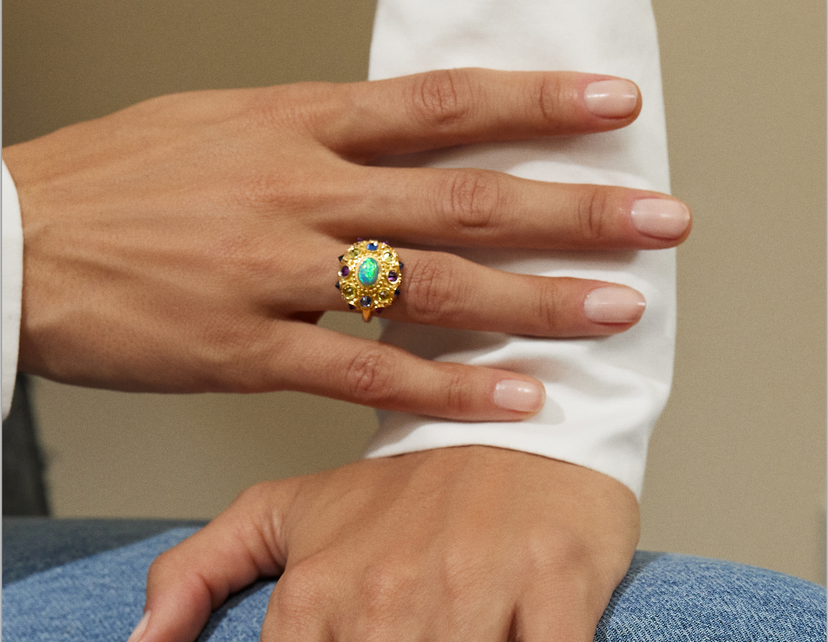 A closeup of the hands of a model showcasing AnaKatarina's statement piece: the Blue Valentine Ring. Crafted from 18k yellow gold and adorned with a Lighting Ridge black opal and vibrant peridots, amethysts, and blue sapphires. With spiritual properties a