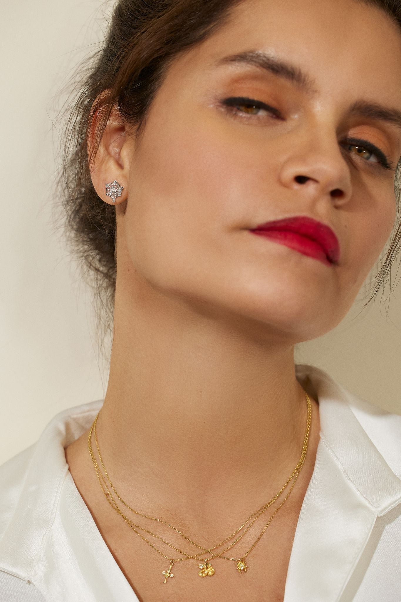A modeling wearing three 18k yellow gold necklaces from Ana-Katarina fine jewelry’s ‘Impeccable Words’ collection: the North Star Cross, a serpent, and a spider - as well as a spider web stud earring in white gold. 